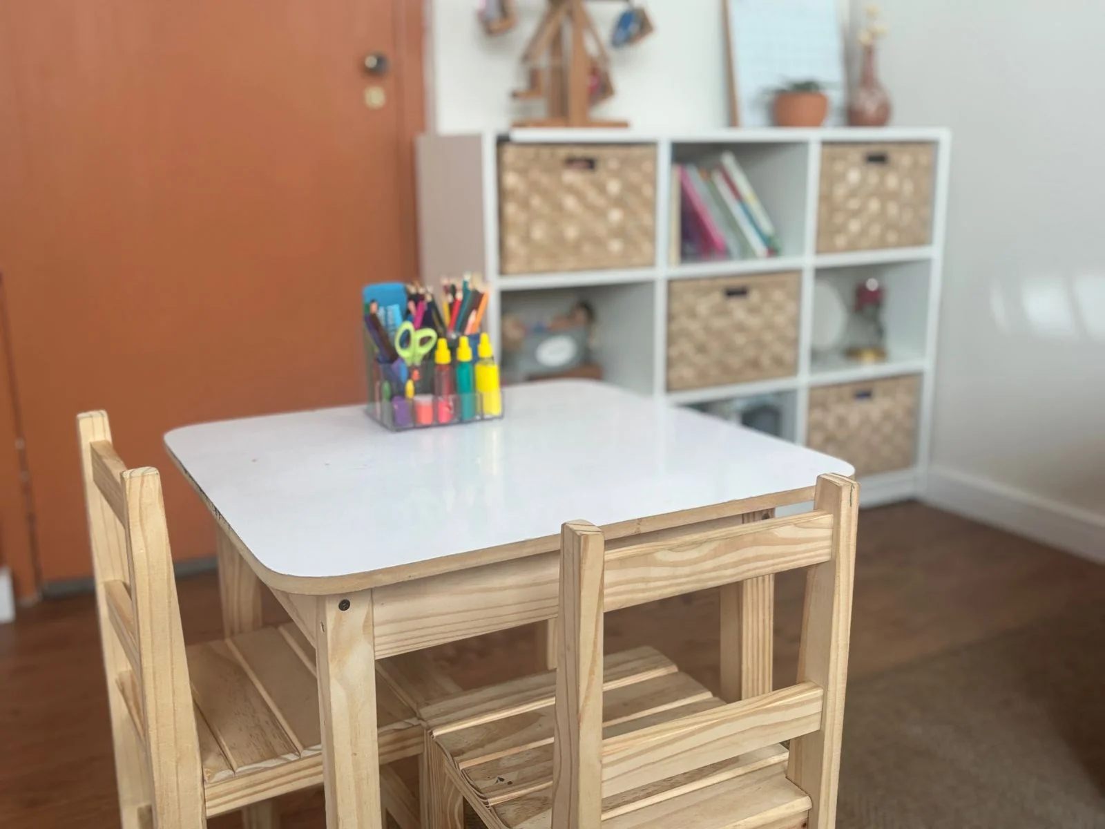 Mesa de atividades infantil com lápis de cor, ao lado de uma estante com livros e brinquedos